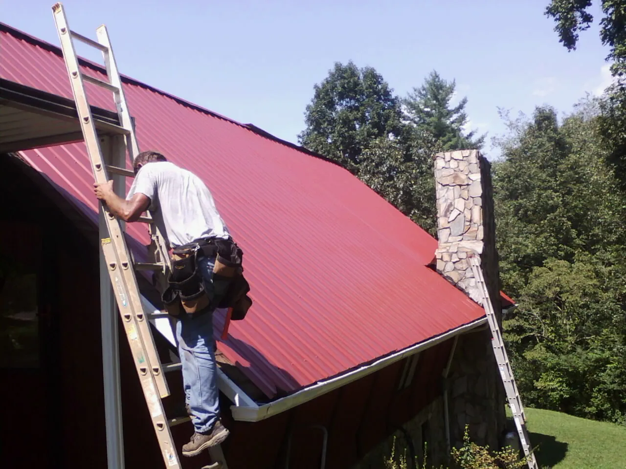 Roofer on a ladder beside a red metal roof and stone chimney — Soffit & Fascia Repair by Roofing Brothers in Taylor Ridge